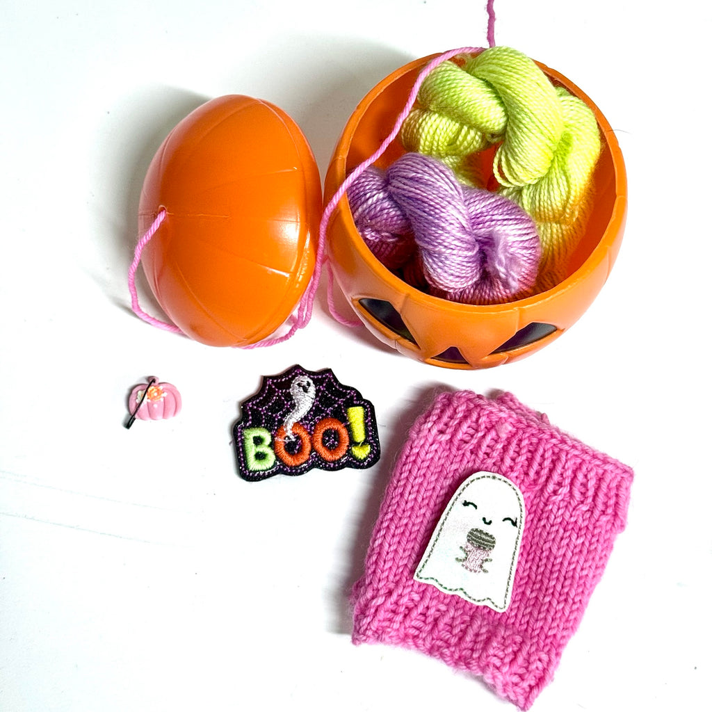 Halloween Mug Cozy Kit with pumpkin yarn bowl, mini skeins, stitch marker, embroidered patch, and pink knitted mug cozy